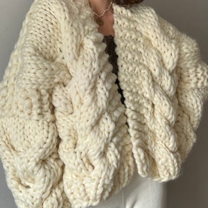 May include: A chunky knit cardigan in a cream color with a cable knit pattern. The cardigan is open in the front and has a relaxed fit.