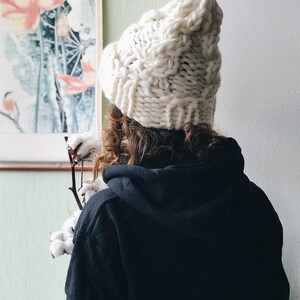 May include: A person wearing a white chunky knit beanie hat and a black hoodie. The person is standing in front of a wall with a painting of flowers on it.