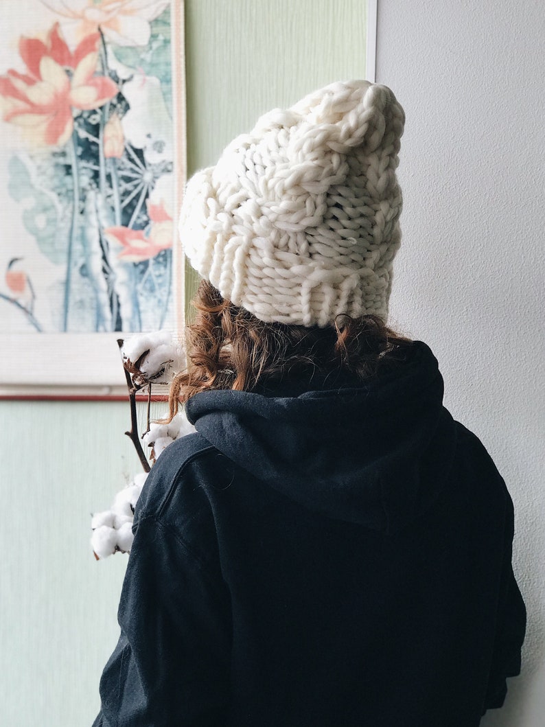 May include: A person wearing a white chunky knit beanie hat and a black hoodie. The person is standing in front of a wall with a painting of flowers on it.
