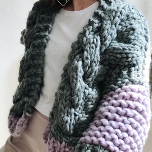 May include: A chunky knit cardigan with long sleeves. The cardigan is a mix of grey and purple yarn. The sleeves are a solid purple colour, while the body of the cardigan is a grey colour with a cable knit pattern.