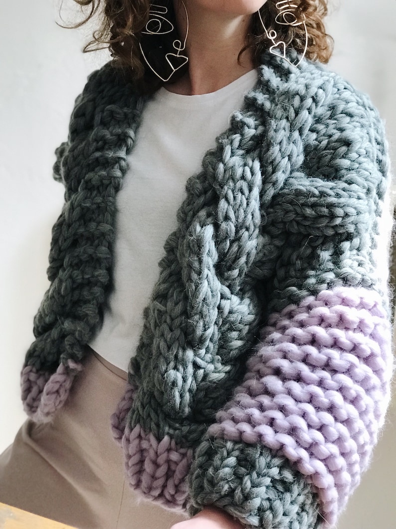 May include: A chunky knit cardigan with long sleeves. The cardigan is a mix of grey and purple yarn. The sleeves are a solid purple colour, while the body of the cardigan is a grey colour with a cable knit pattern.