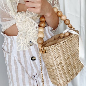 May include: A woven straw tote bag with a wooden bead handle and a gold chain strap. The bag is light brown and has a white ribbon attached to the handle.