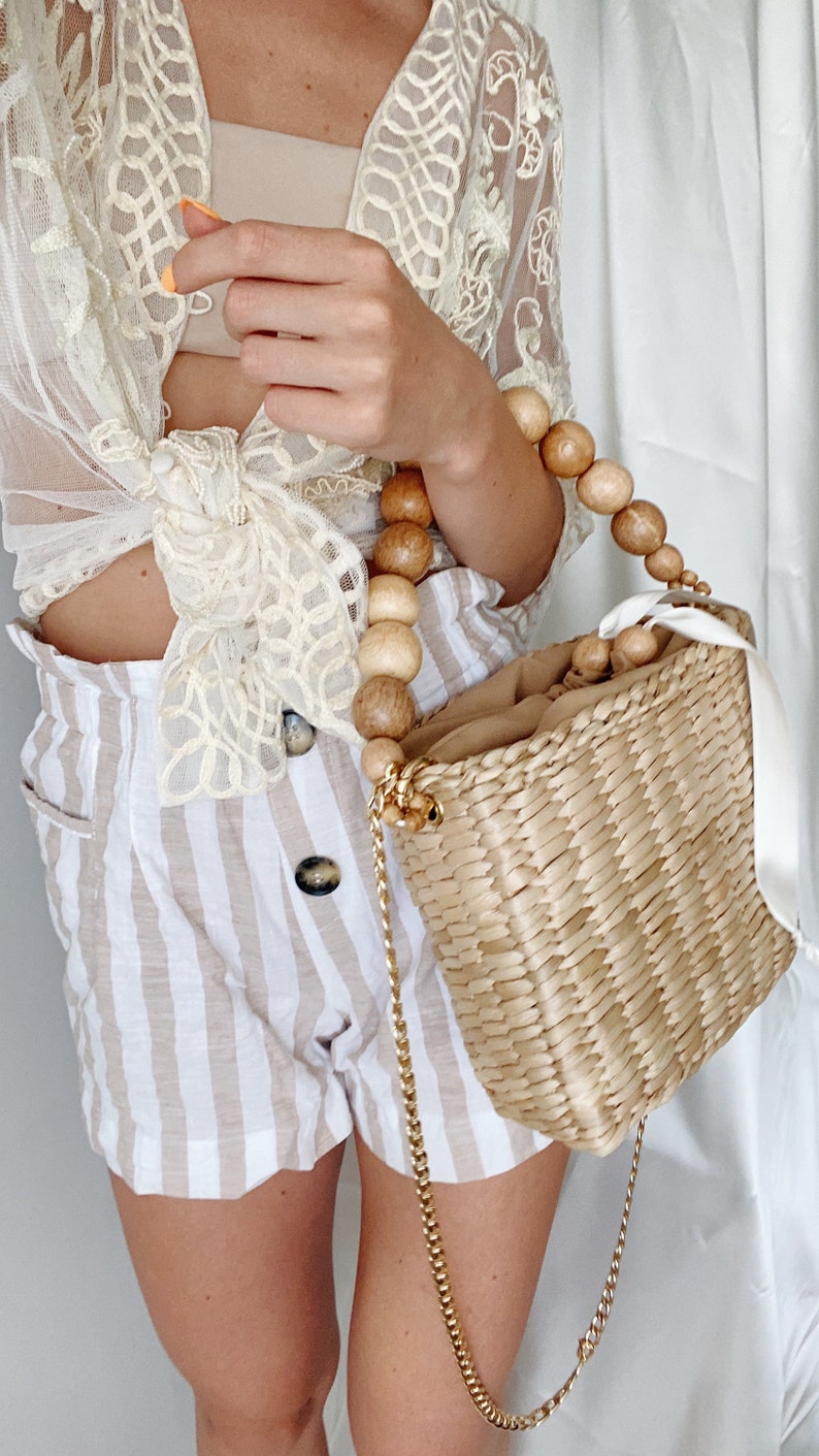 May include: A woven straw tote bag with a wooden bead handle and a gold chain strap. The bag is light brown and has a white ribbon attached to the handle.