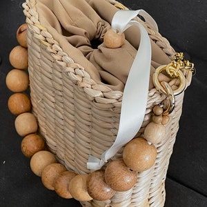 May include: A woven straw bag with a white ribbon handle and a wooden bead chain. The bag has a beige fabric lining and a gold chain strap.