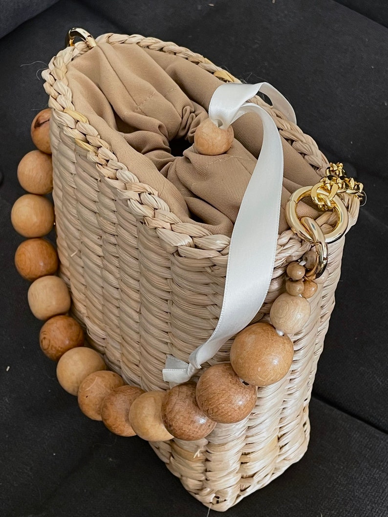 May include: A woven straw bag with a white ribbon handle and a wooden bead chain. The bag has a beige fabric lining and a gold chain strap.
