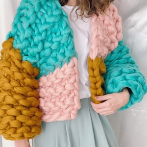 Pode incluir: Um casaco de malha grossa com um design de blocos de cores em turquesa, rosa e amarelo mostarda. O casaco tem um caimento solto e &eacute; feito com um fio grosso.