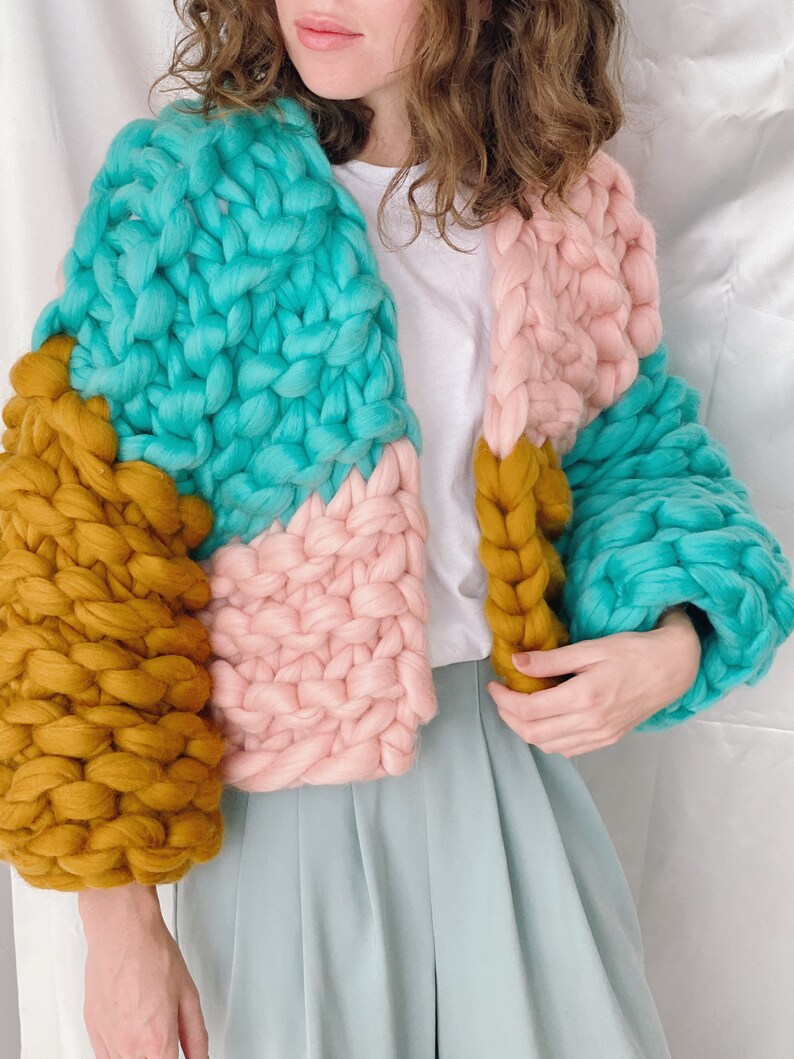 Pode incluir: Um casaco de malha grossa com um design de blocos de cores em turquesa, rosa e amarelo mostarda. O casaco tem um caimento solto e &eacute; feito com um fio grosso.