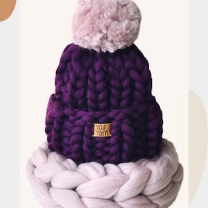 May include: A purple knitted beanie with a fluffy pink pom pom and a brown leather tag that says "Your Yarn". The beanie is sitting on top of a thick, braided, light purple and white yarn.