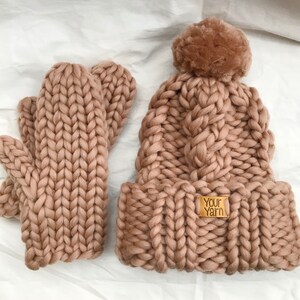 May include: A chunky knit hat and matching mittens in a light brown color. The hat has a fluffy pom pom on top and the mittens are fingerless. The hat has a label that says "Your Yarn".