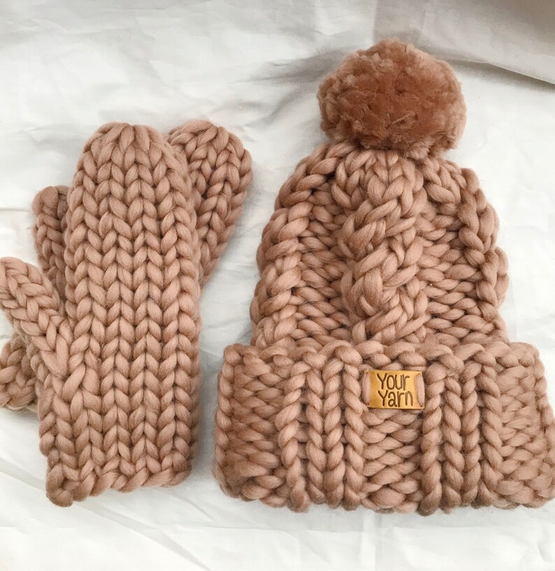 May include: A chunky knit hat and matching mittens in a light brown color. The hat has a fluffy pom pom on top and the mittens are fingerless. The hat has a label that says "Your Yarn".