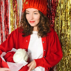 May include: A woman in a red chunky knit hat and cardigan, holding a red box with red and white yarn. The hat has a small tag with the text "YOUR STORY". The background is decorated with red and gold tinsel, creating a festive atmosphere.