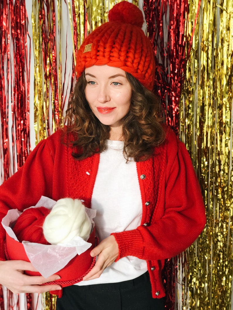 May include: A woman in a red chunky knit hat and cardigan, holding a red box with red and white yarn. The hat has a small tag with the text "YOUR STORY". The background is decorated with red and gold tinsel, creating a festive atmosphere.