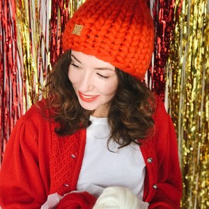 May include: A vibrant orange, chunky knit hat with a large pom-pom. The hat has a small tag that reads "Your Yarn". The person in the image is wearing a red cardigan and holding a box with red and white yarn.