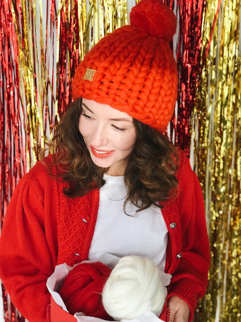 May include: A vibrant orange, chunky knit hat with a large pom-pom. The hat has a small tag that reads "Your Yarn". The person in the image is wearing a red cardigan and holding a box with red and white yarn.