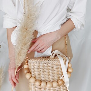 May include: A woven straw bag with a white ribbon and wooden bead accents. The bag is worn as a crossbody bag with a gold chain strap.