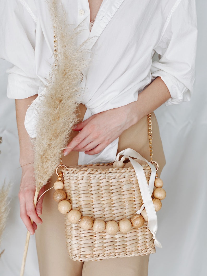 May include: A woven straw bag with a white ribbon and wooden bead accents. The bag is worn as a crossbody bag with a gold chain strap.
