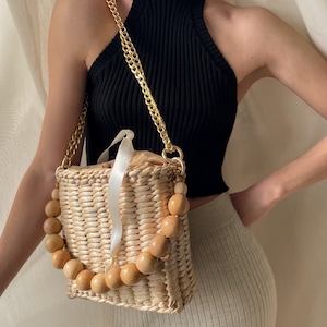 May include: A woven straw bag with a wooden bead handle and a gold chain strap. The bag is light brown and has a white ribbon detail.