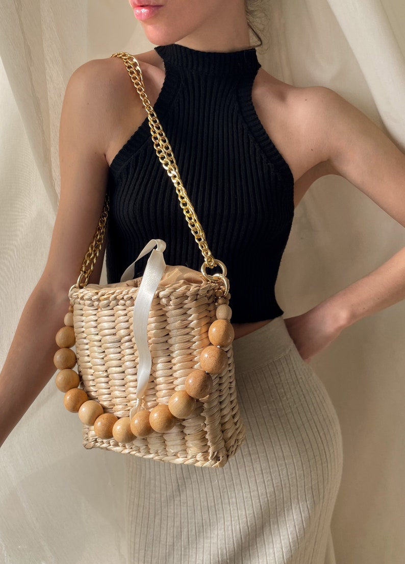 May include: A woven straw bag with a wooden bead handle and a gold chain strap. The bag is light brown and has a white ribbon detail.