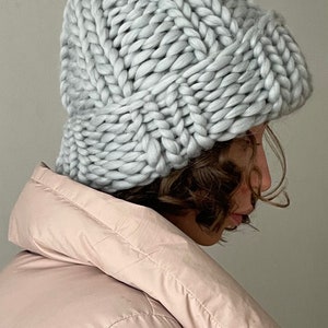 May include: A grey knitted slouchy beanie hat with a thick cable knit pattern. The hat is worn by a person with brown hair and a pink puffer jacket.
