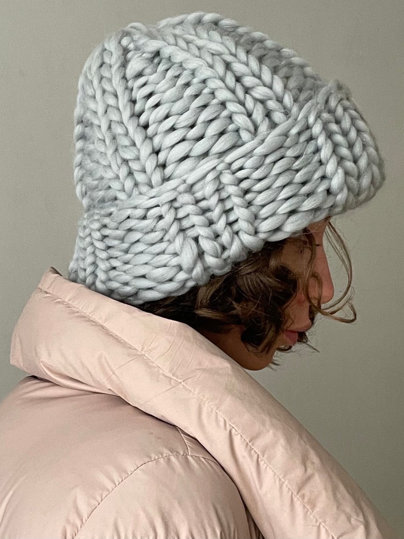 May include: A grey knitted slouchy beanie hat with a thick cable knit pattern. The hat is worn by a person with brown hair and a pink puffer jacket.