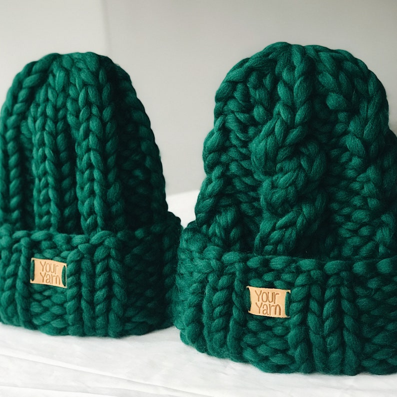 May include: Two dark green knitted beanies with a brown leather tag that says "Your Yarn" on each.