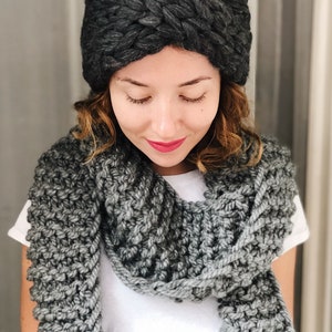 May include: A woman wearing a gray knit headband and a gray knit scarf. The headband is a thick, chunky knit with a braided design. The scarf is a simple knit with a textured pattern.