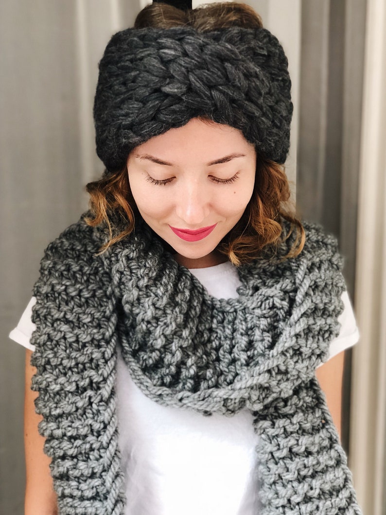 May include: A woman wearing a gray knit headband and a gray knit scarf. The headband is a thick, chunky knit with a braided design. The scarf is a simple knit with a textured pattern.