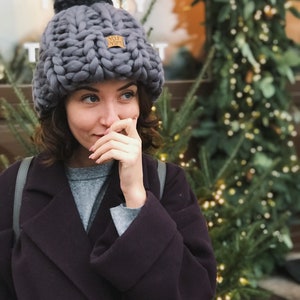 May include: A woman wearing a gray knitted beanie with a pom pom and a brown leather patch that says "Your Yarn". She is wearing a gray sweater and a dark purple coat.