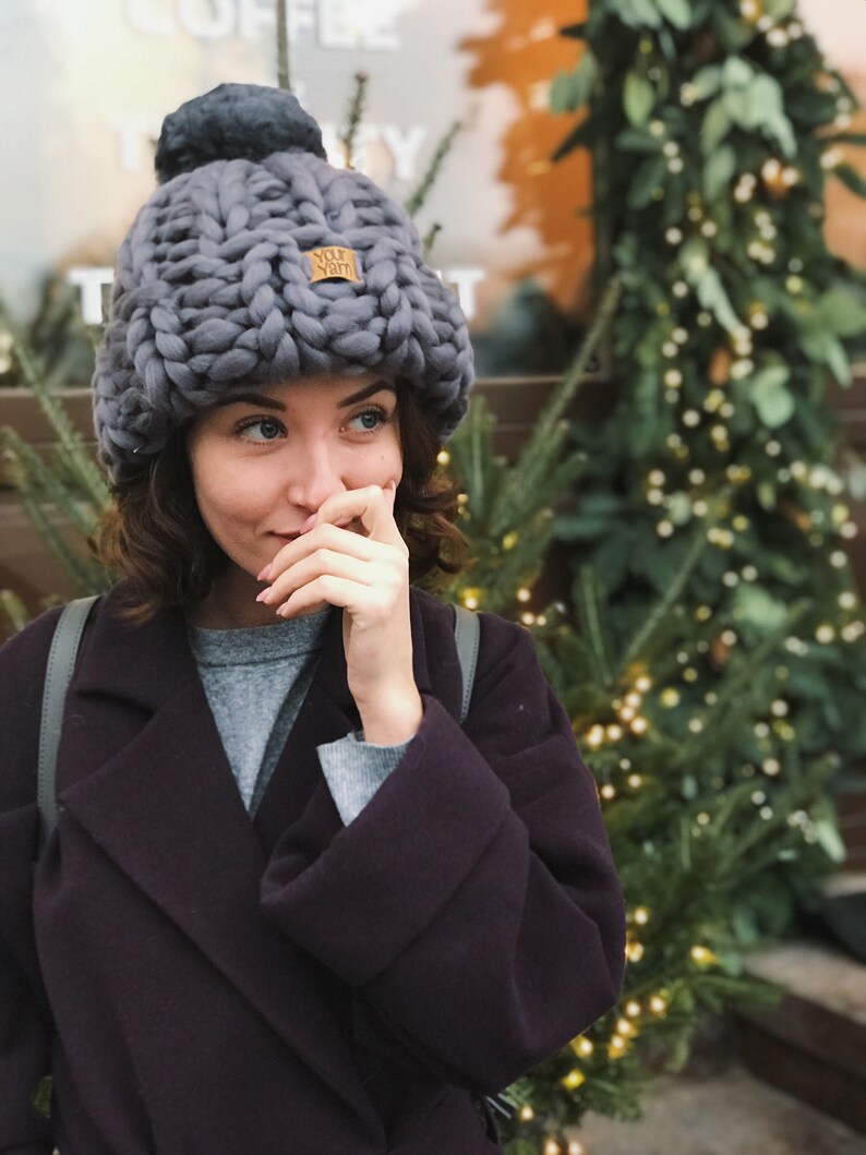 May include: A woman wearing a gray knitted beanie with a pom pom and a brown leather patch that says "Your Yarn". She is wearing a gray sweater and a dark purple coat.