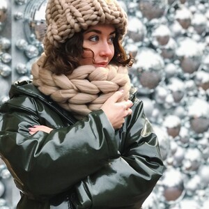 May include: A woman wearing a beige knitted hat and scarf with a green, shiny jacket. The woman is standing in front of a wall of white and silver ornaments.