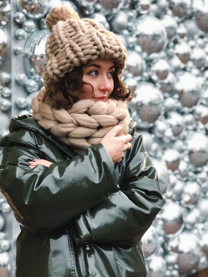 May include: A woman wearing a beige knitted hat and scarf with a green, shiny jacket. The woman is standing in front of a wall of white and silver ornaments.