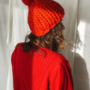 May include: A bright orange knitted beanie hat with a large pom-pom. The chunky knit hat is made of thick yarn and has a small, rectangular tag on the side. The person in the image is wearing a matching red sweater.