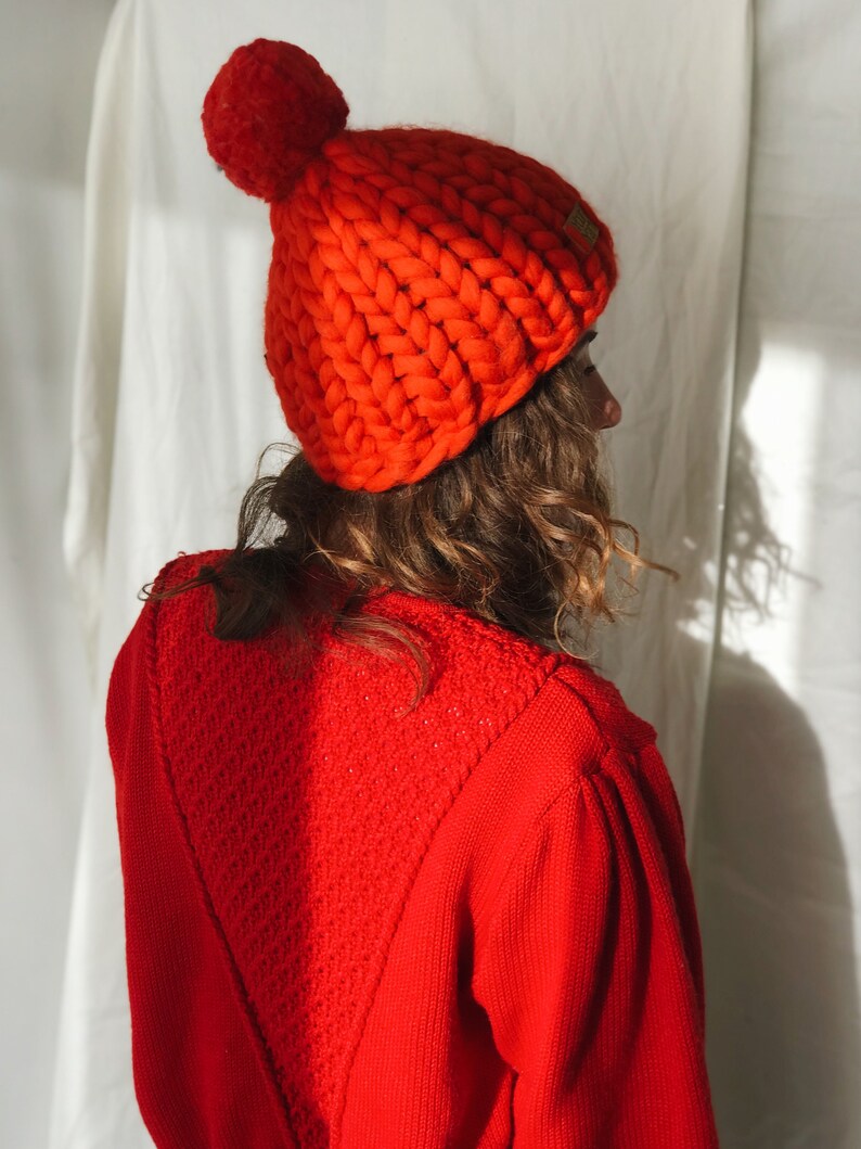 May include: A bright orange knitted beanie hat with a large pom-pom. The chunky knit hat is made of thick yarn and has a small, rectangular tag on the side. The person in the image is wearing a matching red sweater.