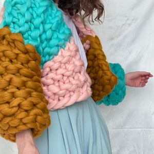 Pode incluir: Um casaco de malha grossa com um design de blocos de cores em turquesa, rosa e marrom. O casaco tem um caimento solto e mangas compridas.