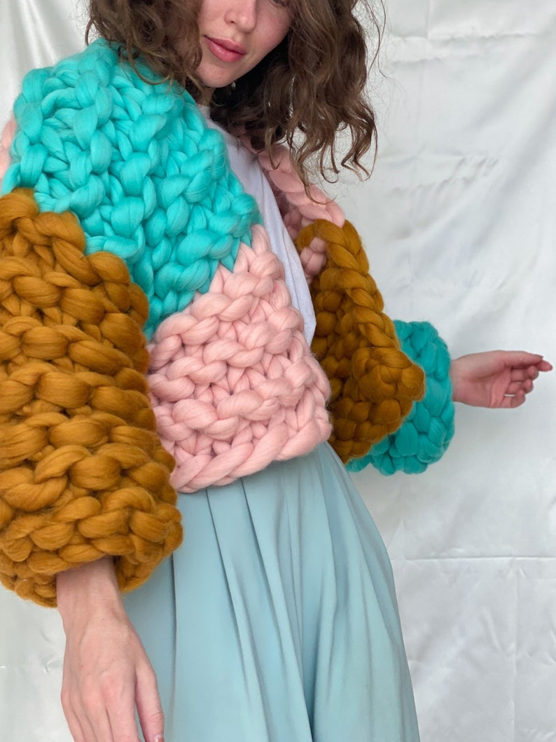 Pode incluir: Um casaco de malha grossa com um design de blocos de cores em turquesa, rosa e marrom. O casaco tem um caimento solto e mangas compridas.