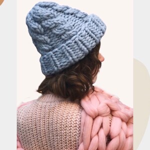 May include: A light blue knitted beanie with a chunky cable knit pattern. The beanie is worn on a person's head, with their back to the camera. The person is wearing a pink knitted sweater.