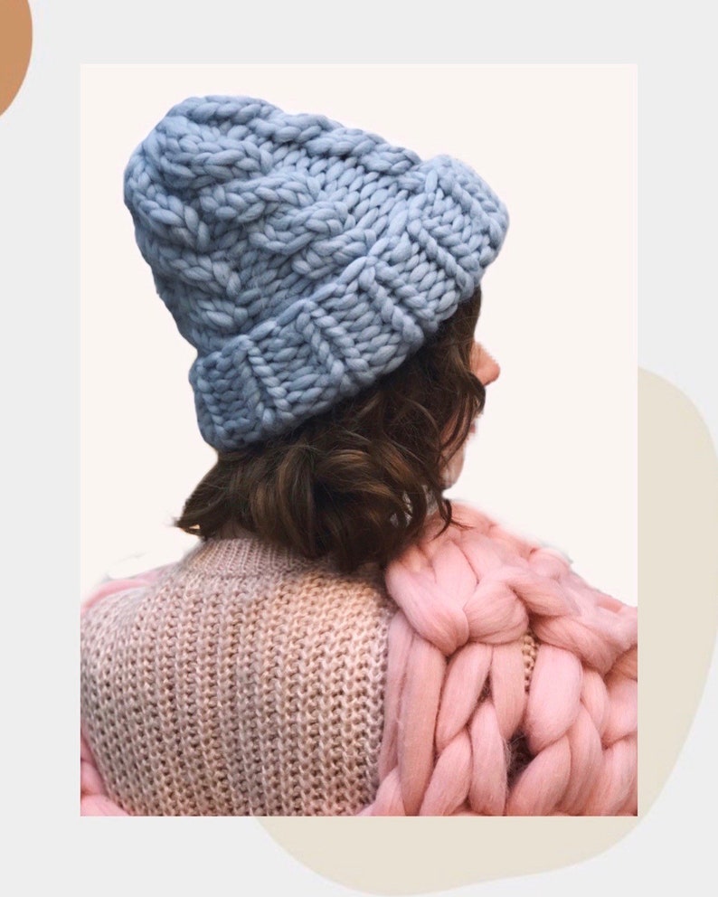 May include: A light blue knitted beanie with a chunky cable knit pattern. The beanie is worn on a person's head, with their back to the camera. The person is wearing a pink knitted sweater.