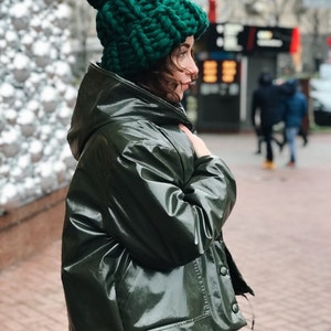 May include: A woman wearing a green hooded jacket and a green knit beanie with a pom pom. The jacket is shiny and has a zipper and buttons.