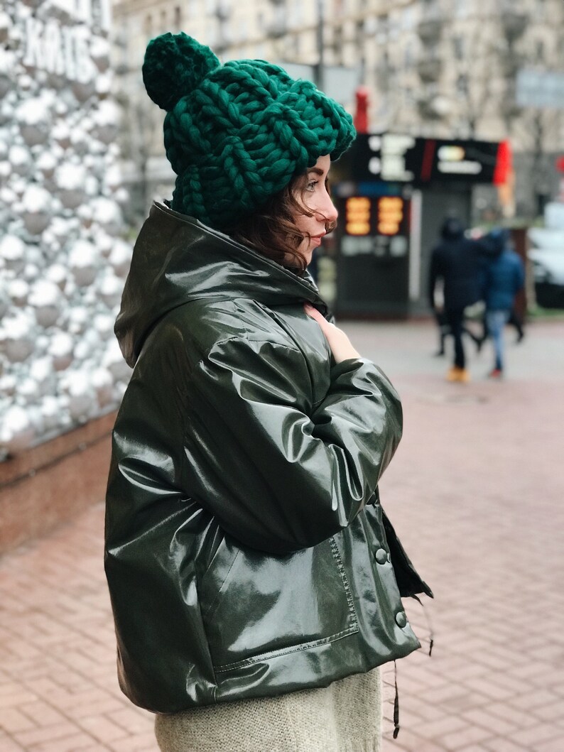 May include: A woman wearing a green hooded jacket and a green knit beanie with a pom pom. The jacket is shiny and has a zipper and buttons.