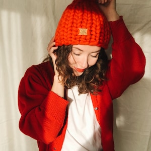 May include: A vibrant red, chunky knit beanie hat with a large pom-pom. The hat has a small tag that reads "Your Yarn". The person in the image is wearing a red cardigan over a white t-shirt.