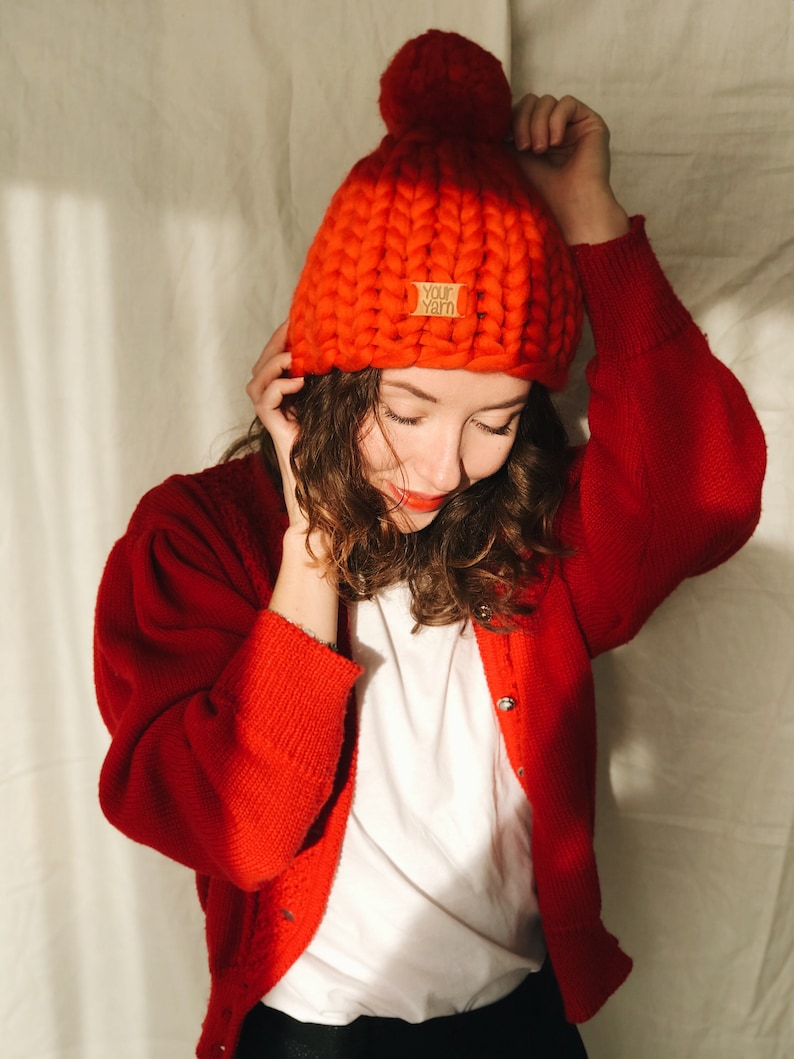 May include: A vibrant red, chunky knit beanie hat with a large pom-pom. The hat has a small tag that reads "Your Yarn". The person in the image is wearing a red cardigan over a white t-shirt.