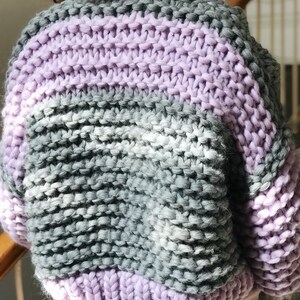 May include: A chunky knit cardigan with stripes of lavender and grey yarn. The cardigan has a relaxed fit and a slightly cropped length.