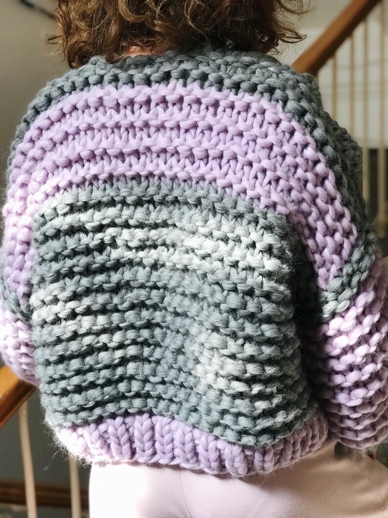 May include: A chunky knit cardigan with stripes of lavender and grey yarn. The cardigan has a relaxed fit and a slightly cropped length.