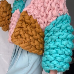 Pode incluir: Um casaco de malha grossa com um design de blocos de cores em rosa, turquesa e amarelo mostarda. O casaco tem um caimento solto e mangas compridas.