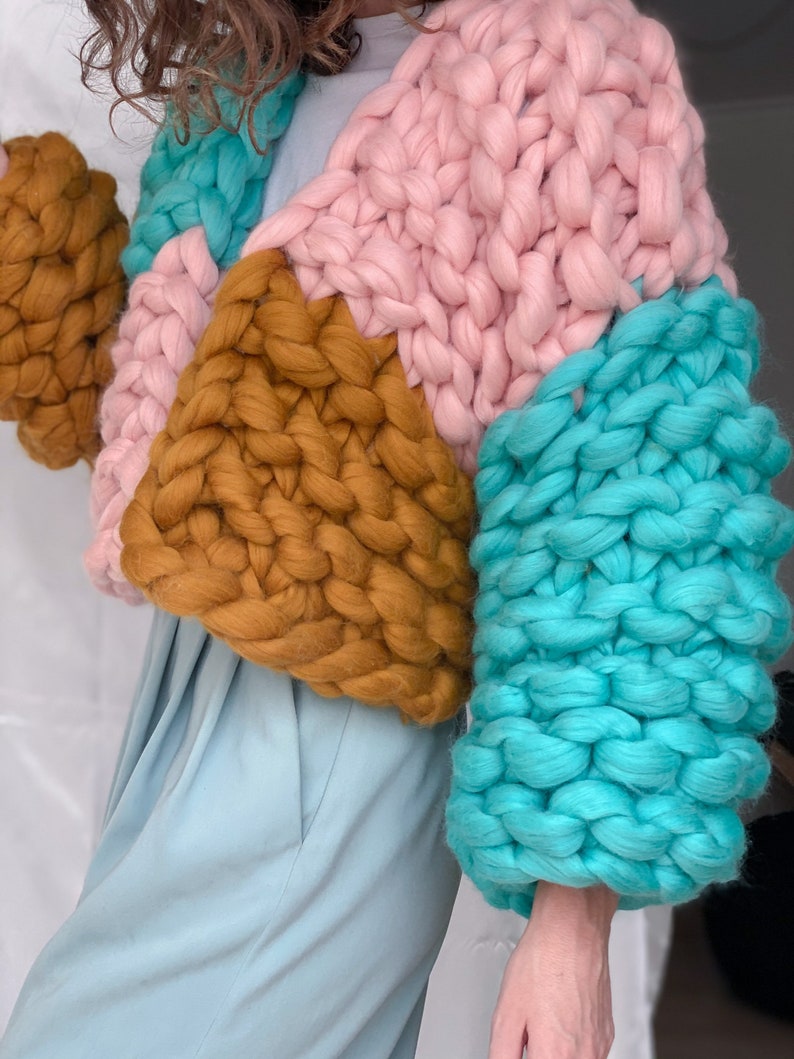 Pode incluir: Um casaco de malha grossa com um design de blocos de cores em rosa, turquesa e amarelo mostarda. O casaco tem um caimento solto e mangas compridas.