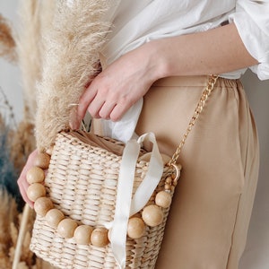 May include: A woven straw bag with a white ribbon and wooden bead accents. The bag has a gold chain strap.
