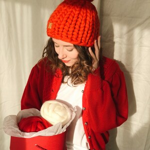 May include: A vibrant red, chunky knit beanie hat with a large pom-pom. The hat features a small gold emblem. A red cardigan and white t-shirt are worn. A red hatbox contains two balls of yarn, one red and one cream.