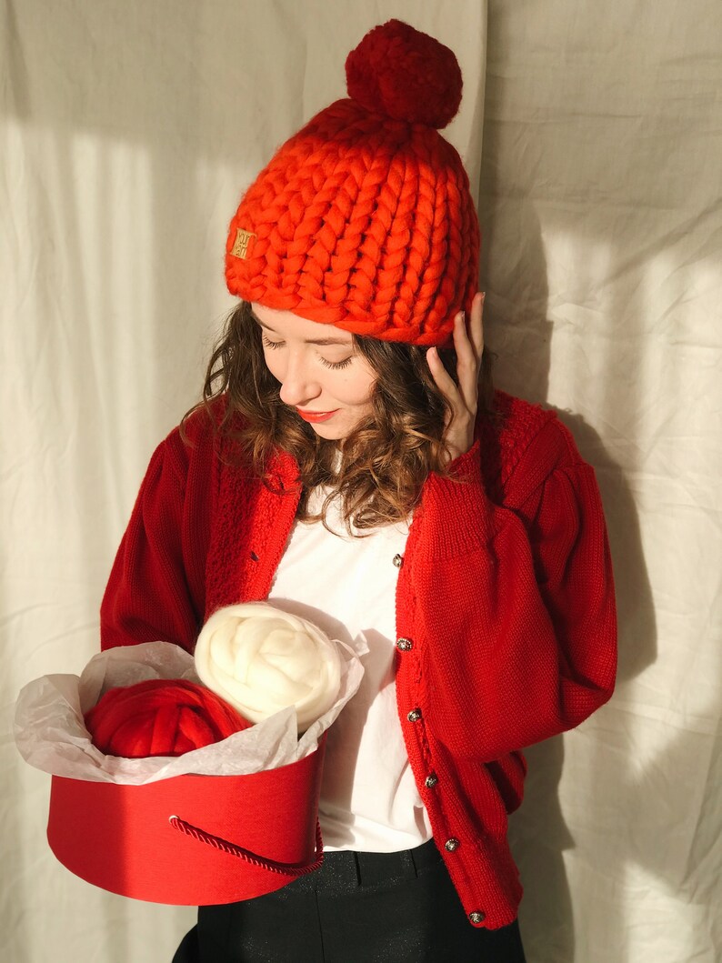 May include: A vibrant red, chunky knit beanie hat with a large pom-pom. The hat features a small gold emblem. A red cardigan and white t-shirt are worn. A red hatbox contains two balls of yarn, one red and one cream.