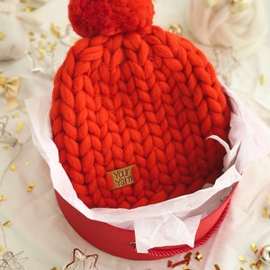 May include: A bright red, chunky-knit beanie hat with a large pom-pom, presented in a matching red gift box lined with white tissue paper. The hat has a small tag that reads "Your Yarn". Festive decorations surround the box.