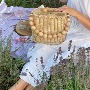May include: A woven straw bag with a wooden bead handle. The bag is light brown and has a natural, rustic look. The bag is sitting on a purple blanket with lavender flowers in the background.